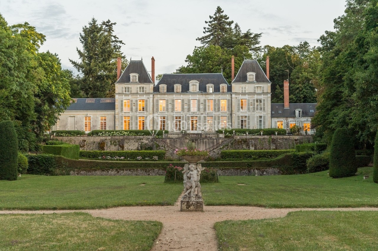 Façade d'un château entouré de jardins verdoyants, avec une statue au premier plan et des arbres en arrière-plan.