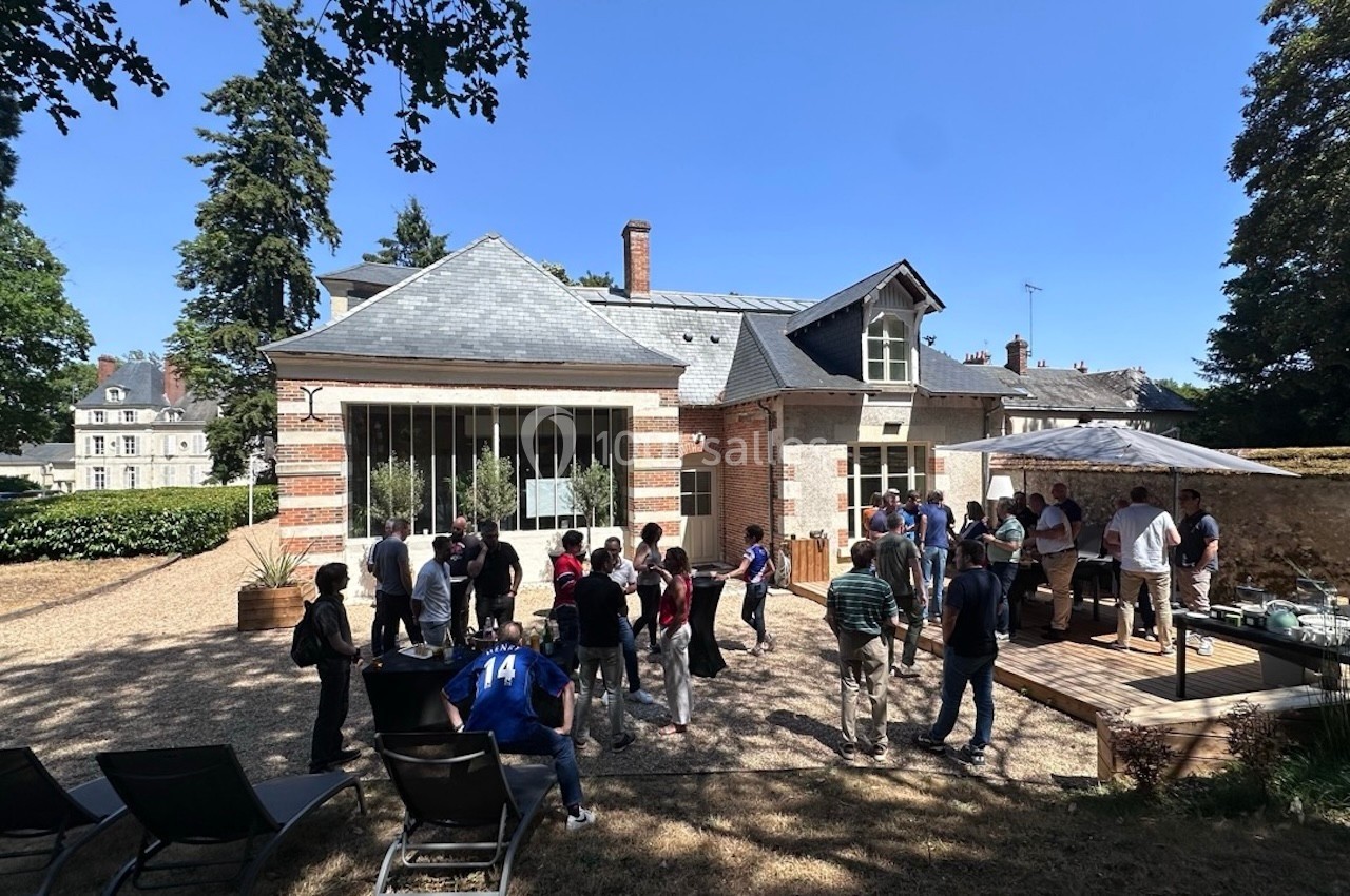 Groupe de personnes rassemblées dans une cour extérieure ensoleillée devant un bâtiment en briques et ardoises.