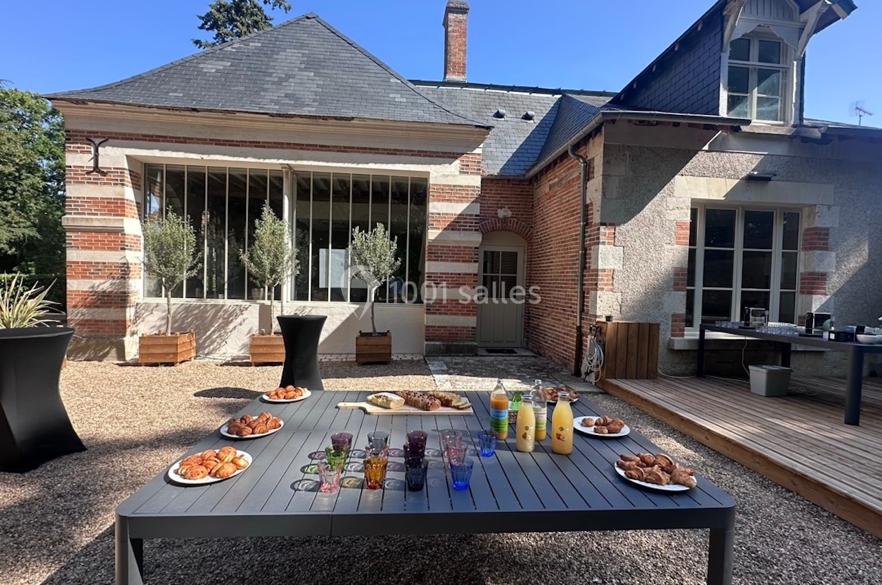 Table avec viennoiseries, jus de fruits et boissons, disposée en extérieur devant une maison en briques et pierres.