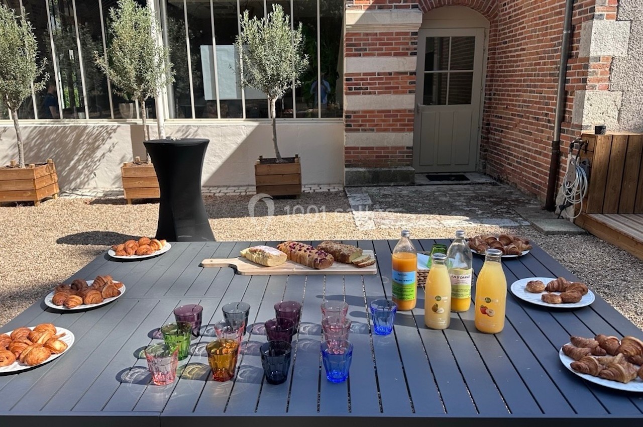 Table extérieure avec viennoiseries, jus de fruits, pain et verres colorés, devant un bâtiment en briques.