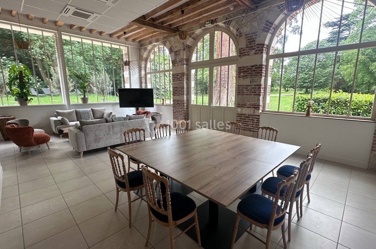 Salle lumineuse avec grande table en bois, chaises, canapés et larges fenêtres donnant sur un jardin verdoyant.