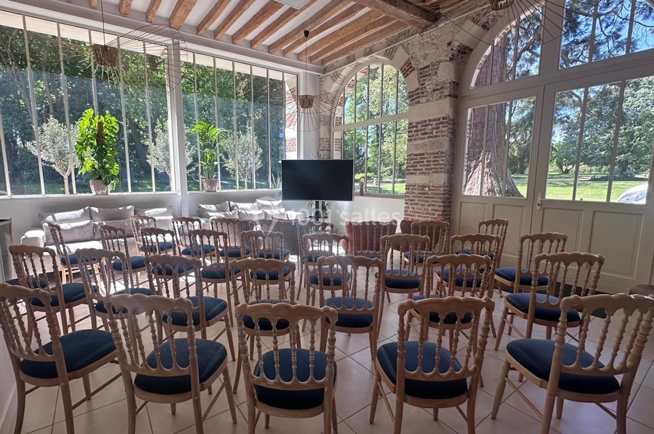 Salle lumineuse avec chaises en bois disposées face à un écran, grandes fenêtres donnant sur un jardin verdoyant.