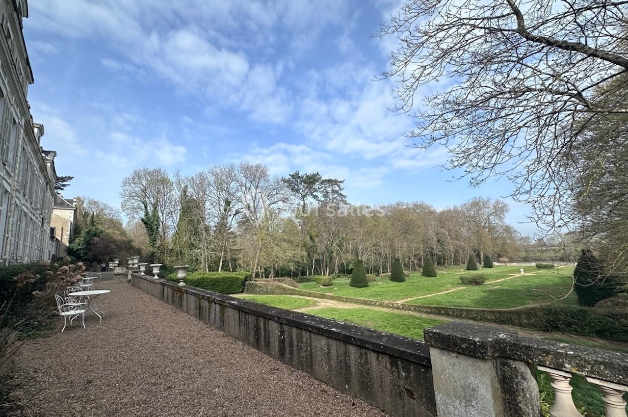 Terrasse en gravier avec tables blanches, surplombant un jardin arboré et des pelouses sous un ciel partiellement nuageux.