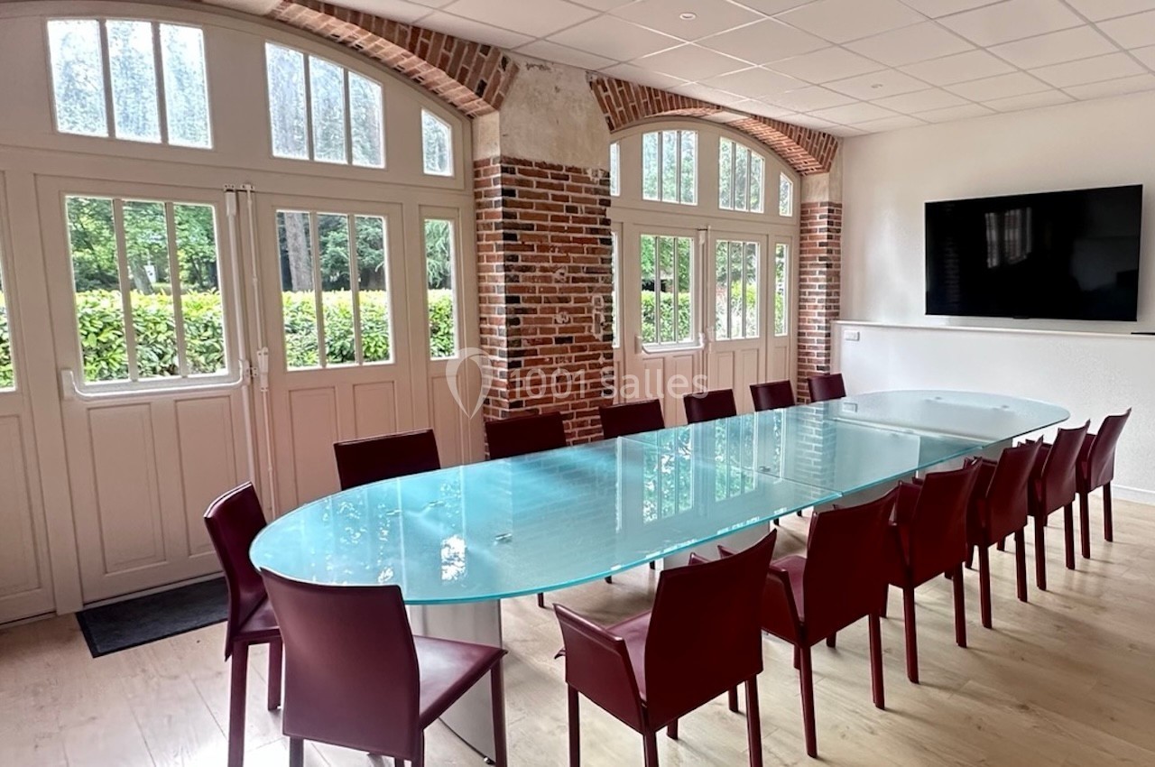 Salle de réunion lumineuse avec une grande table en verre entourée de chaises rouges, fenêtres en arc et mur en briques.