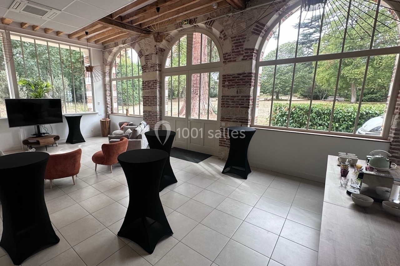 Salle lumineuse avec grandes fenêtres, tables hautes noires, fauteuils et vue sur un espace vert extérieur.