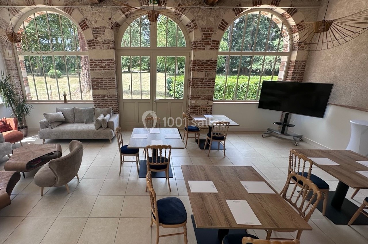 Salle lumineuse avec de grandes baies vitrées, tables en bois, chaises et un écran, donnant sur un jardin verdoyant.