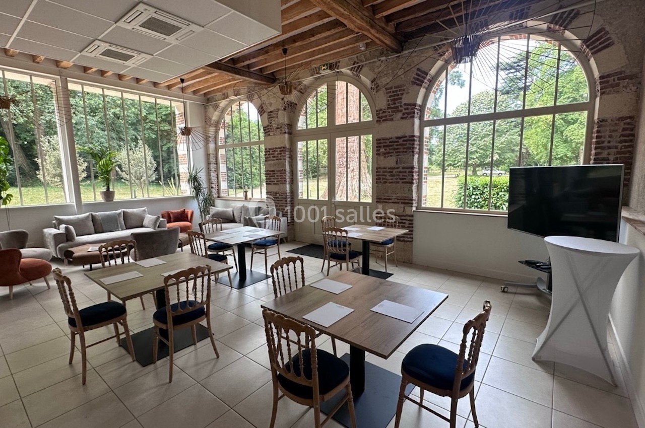 Salle lumineuse avec tables et chaises en bois, grandes fenêtres donnant sur un jardin, et un écran noir sur la droite.