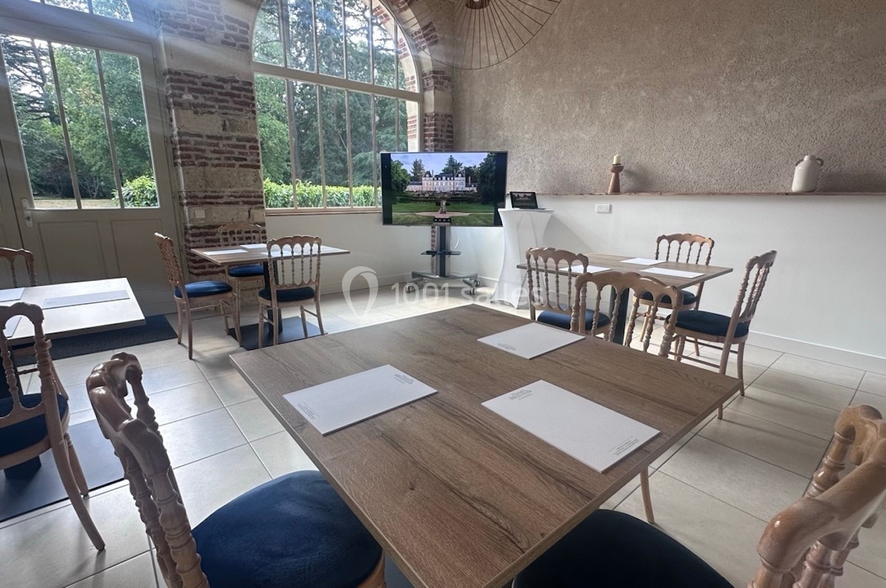 Salle lumineuse avec tables en bois, chaises en bois clair, écran affichant un bâtiment et grandes fenêtres donnant sur un…