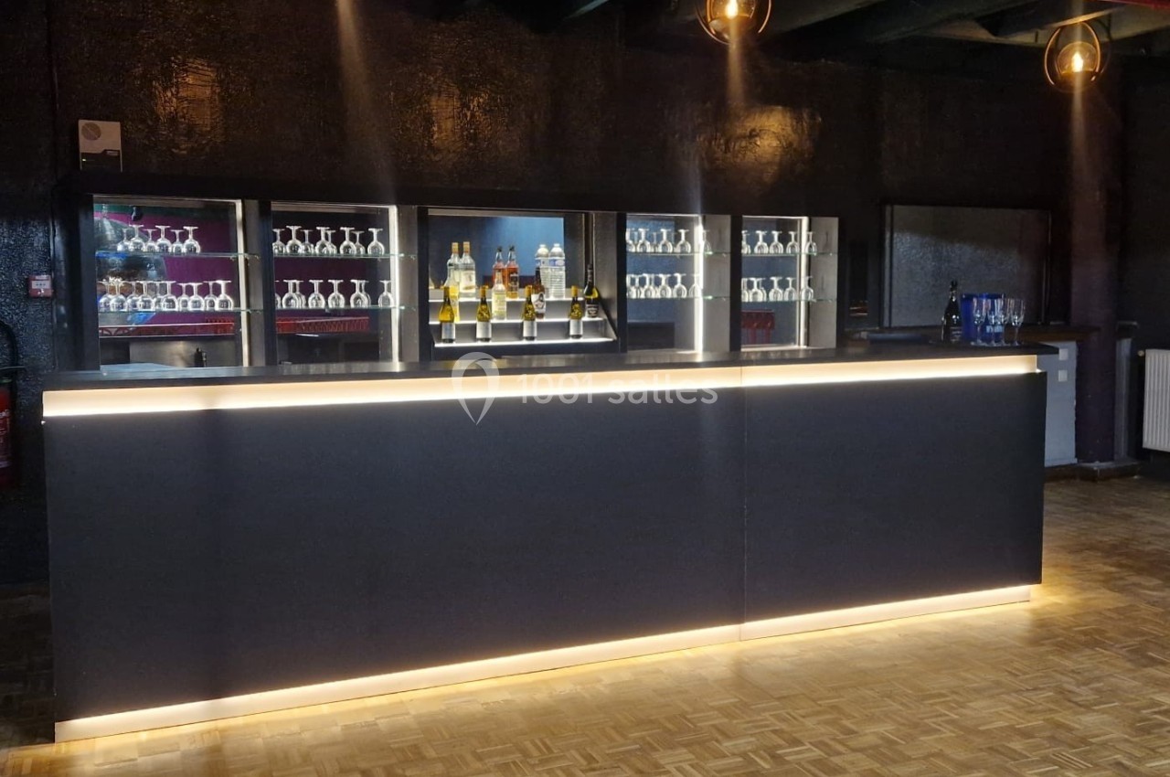 Comptoir de bar moderne éclairé, avec des étagères de bouteilles et de verres en arrière-plan dans un espace sombre.
