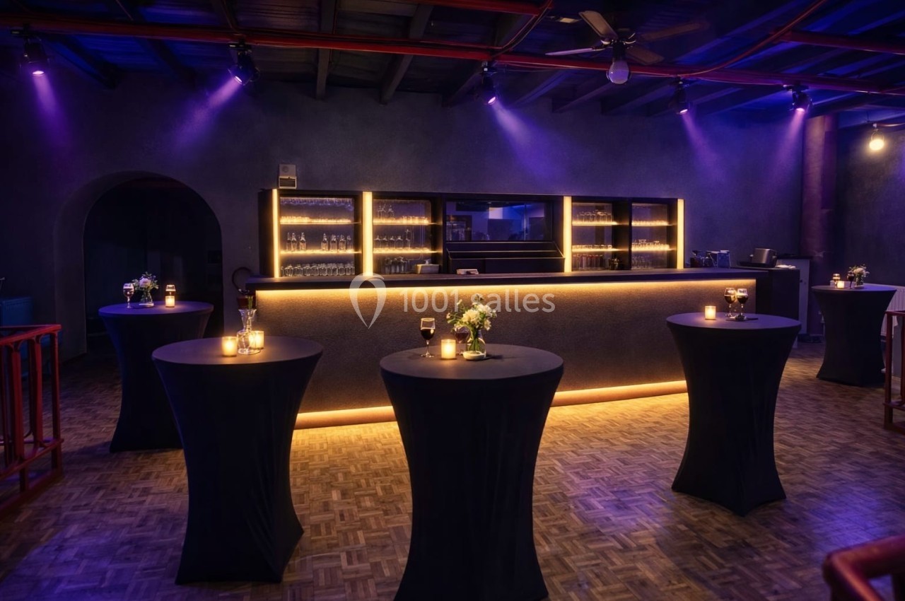 Salle de réception avec tables hautes noires, bougies et fleurs, éclairage tamisé et bar illuminé en arrière-plan.