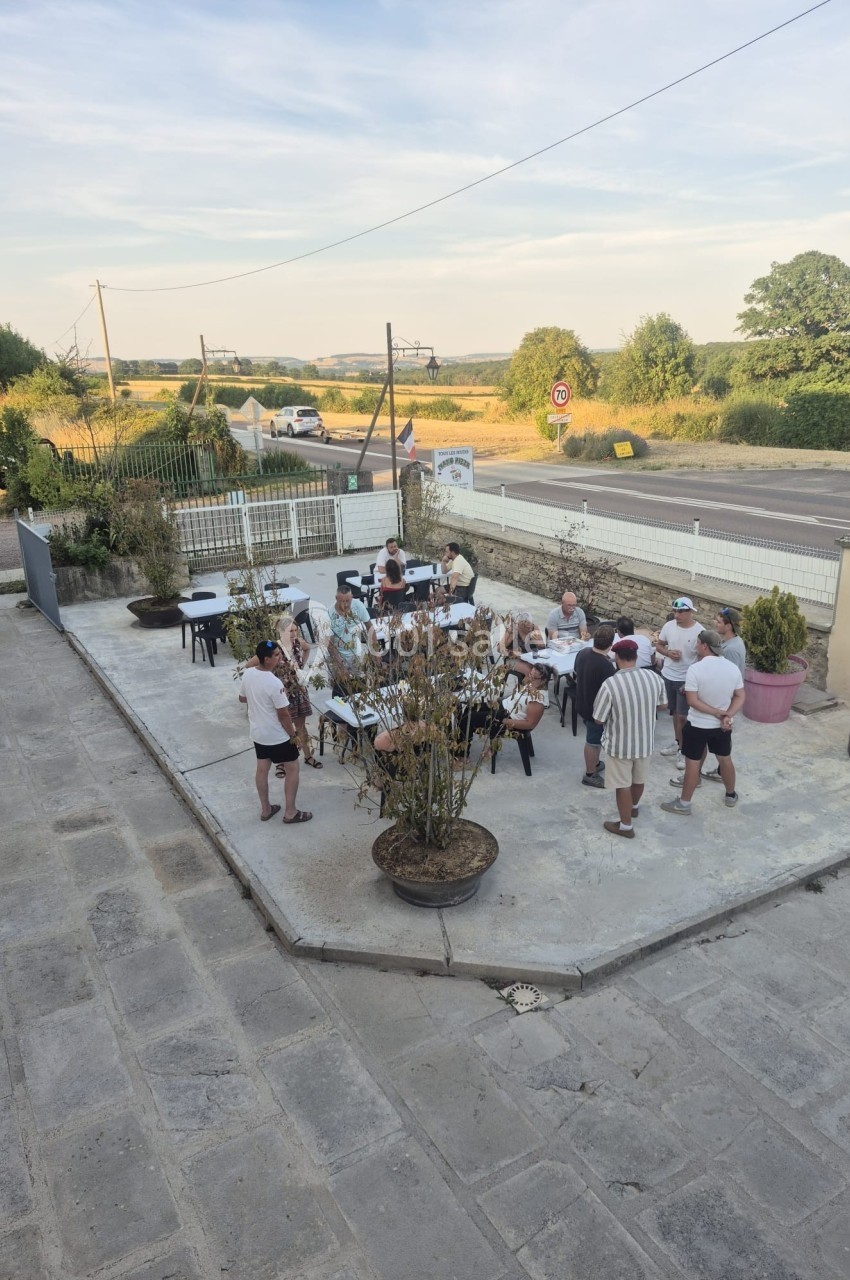 Groupe de personnes discutant autour de tables en extérieur sur une terrasse, avec un paysage rural en arrière-plan.