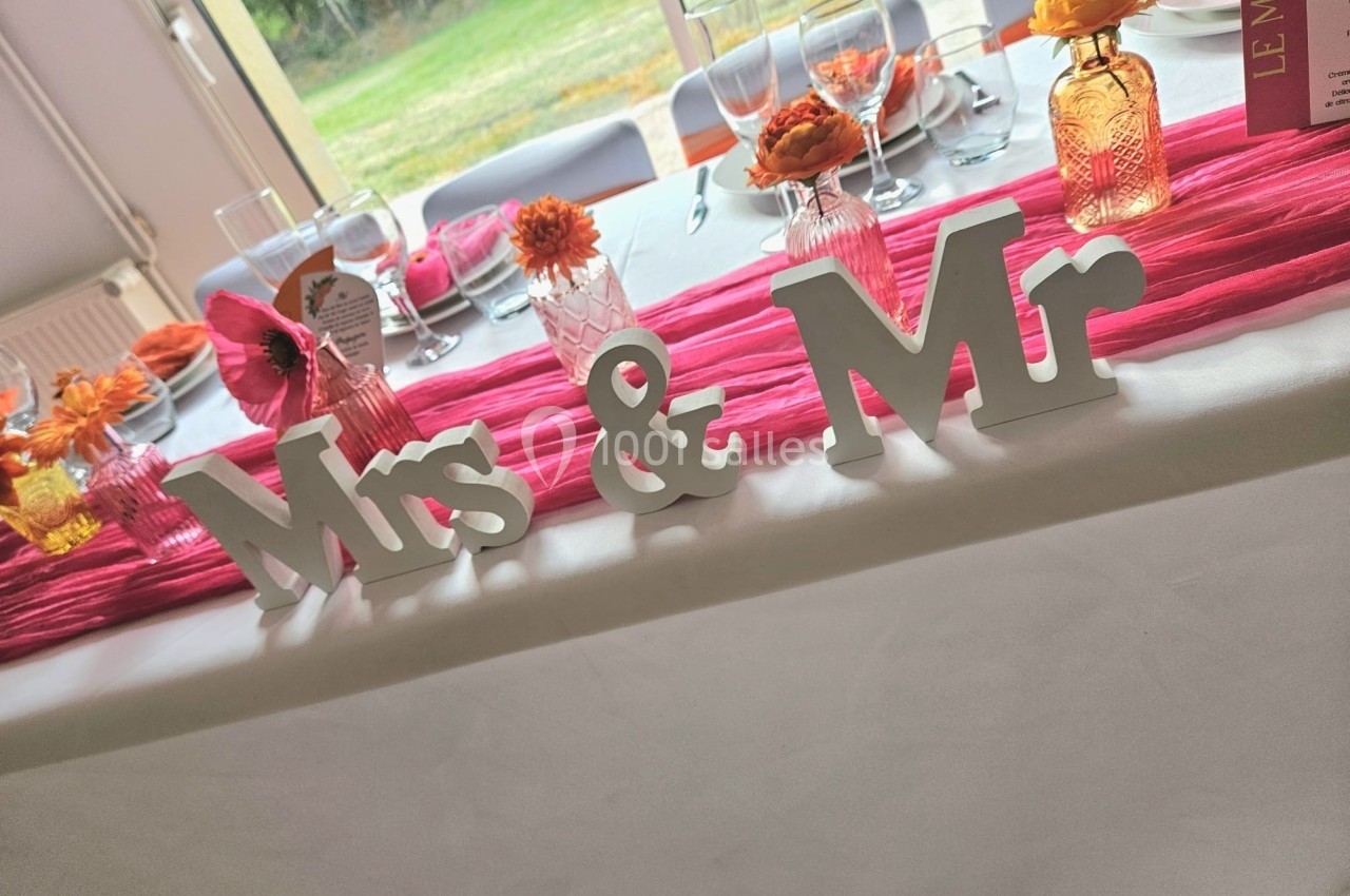 Décoration de table de mariage avec lettres ’Mrs & Mr’, fleurs colorées et nappes roses dans une salle lumineuse.