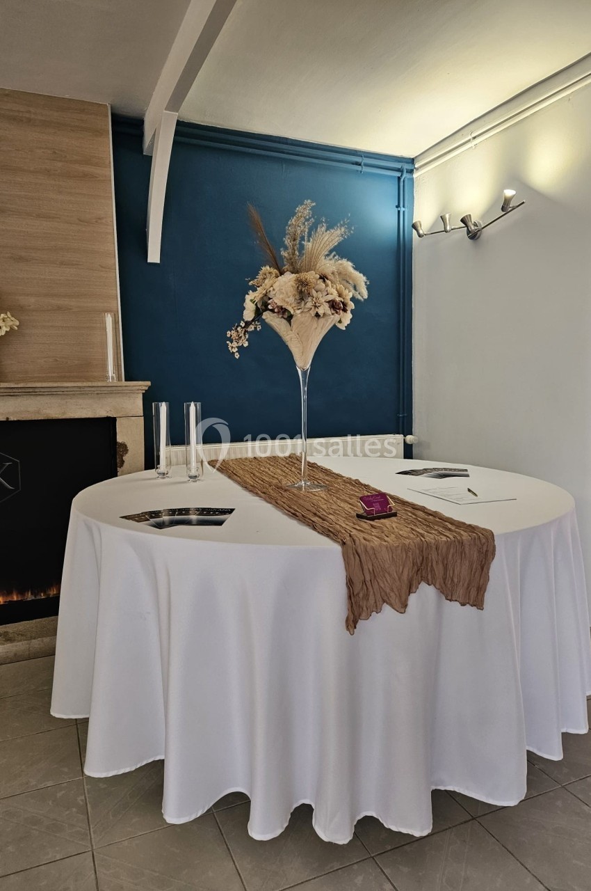 Table ronde décorée avec une nappe blanche, un chemin de table beige et un vase de fleurs séchées au centre.