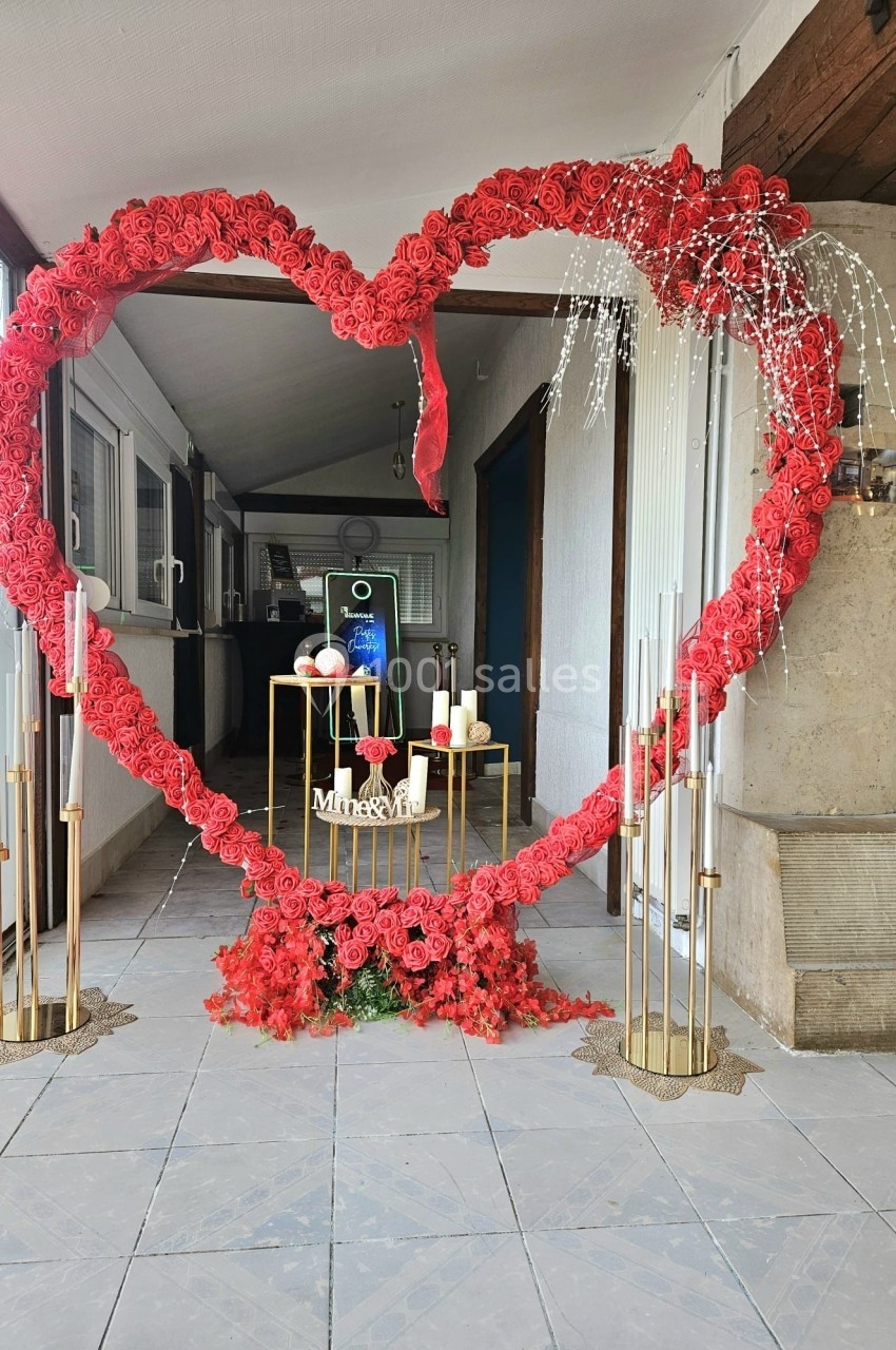 Cœur géant en roses rouges entourant une décoration avec bougies et fleurs dans un espace intérieur lumineux.