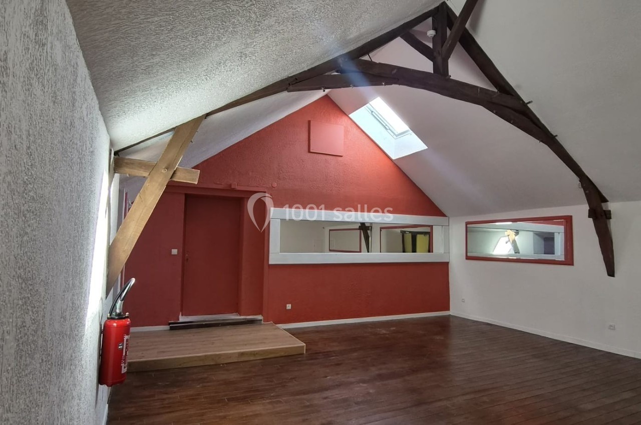 Salle mansardée avec poutres apparentes, murs blancs et rouges, parquet en bois et lucarne laissant entrer la lumière.