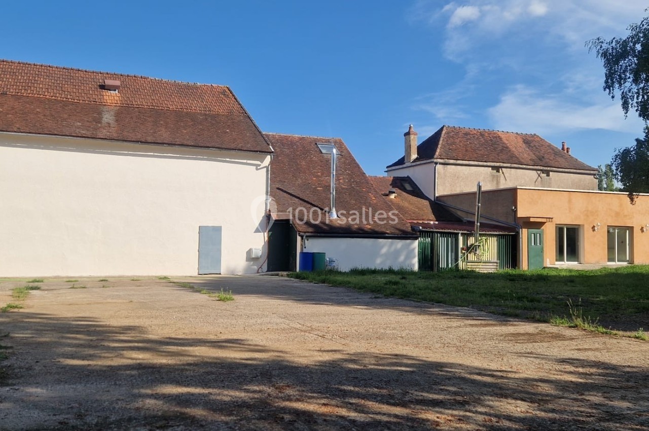 Bâtiments à toits en tuiles entourés de verdure, avec une cour en béton au premier plan sous un ciel dégagé.