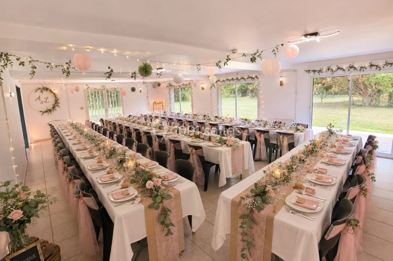 Salle de réception décorée pour un mariage, avec longues tables ornées de fleurs, nappes blanches et rubans roses.