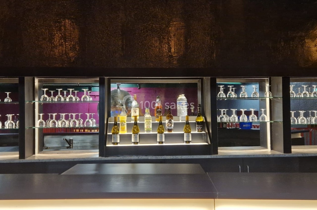 Bar moderne avec étagères en verre illuminées, exposant des verres et des bouteilles d'alcool devant un mur sombre.