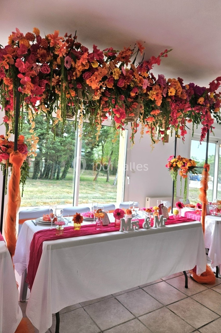 Décoration de table de mariage avec nappes blanches, chemins de table roses et arche florale colorée suspendue.