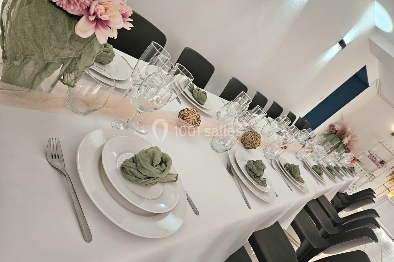 Table dressée avec une nappe blanche, vaisselle élégante, serviettes vertes et décorations florales dans une salle lumineuse.