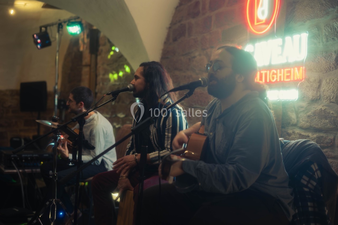 Trois musiciens jouent en live dans un bar, avec des guitares et un micro, devant un mur en pierre et un néon allumé.