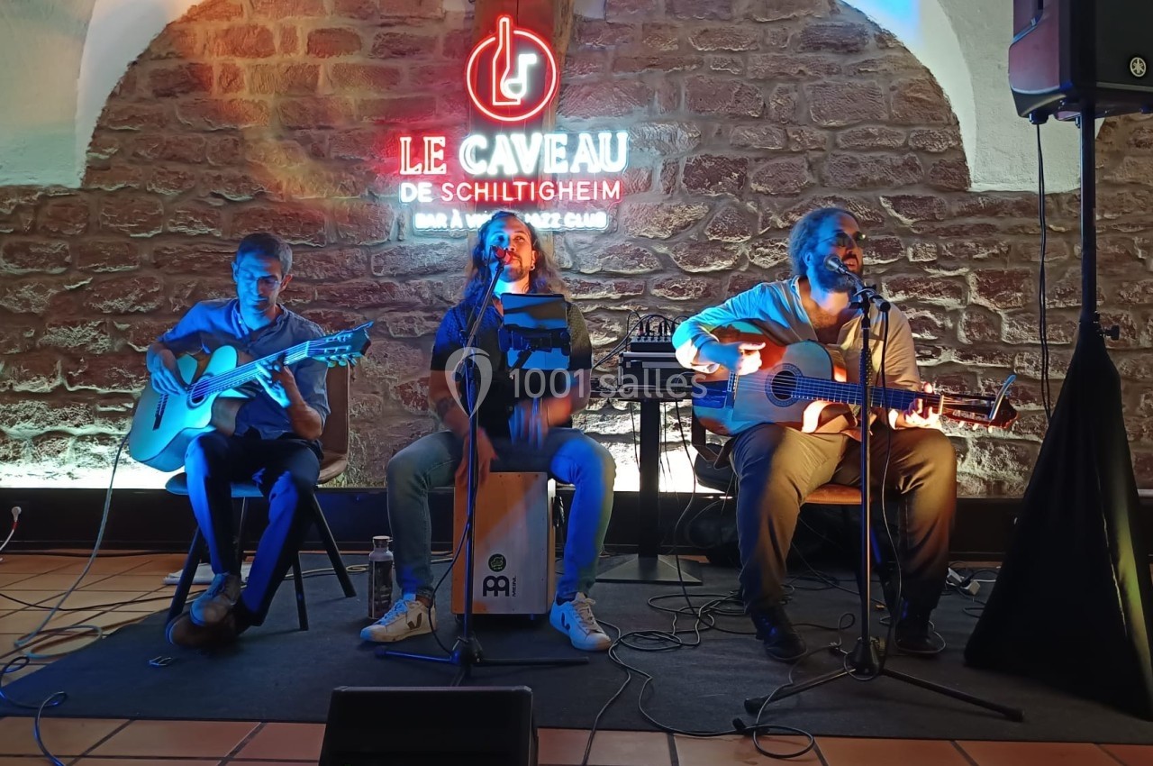Trois musiciens jouent sur scène avec guitares et cajón, devant un mur en pierre éclairé par une enseigne lumineuse.