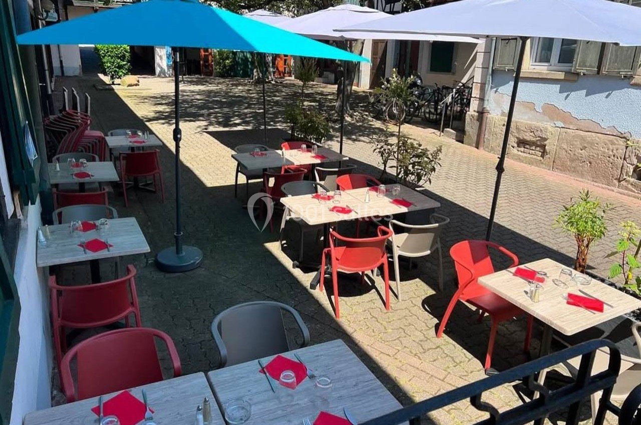 Terrasse extérieure d'un restaurant avec tables dressées, chaises colorées et parasols, située dans une cour pavée.