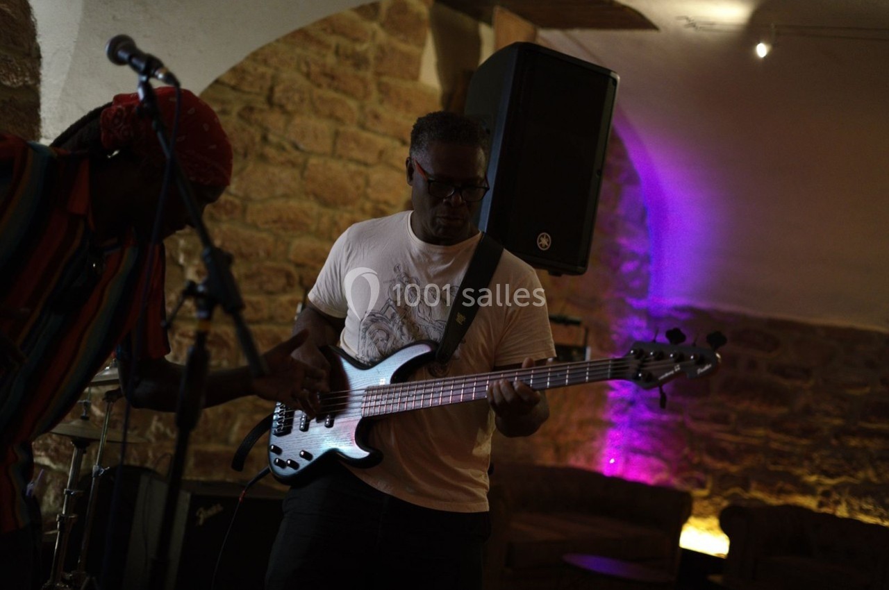 Un musicien joue de la guitare basse dans une salle en pierre éclairée par des lumières violettes.