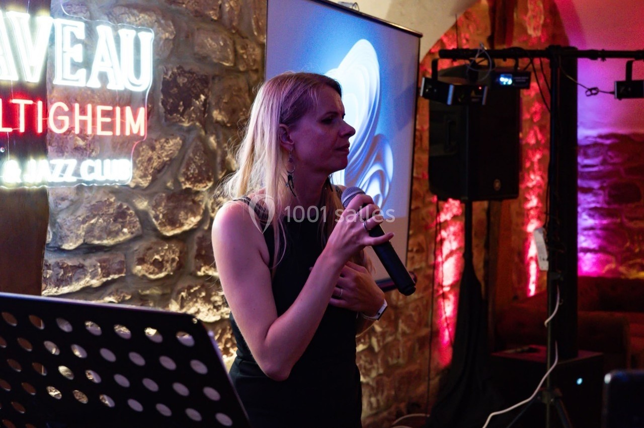 Une femme tenant un micro chante ou parle dans un club, éclairé par des lumières colorées et un écran en arrière-plan.