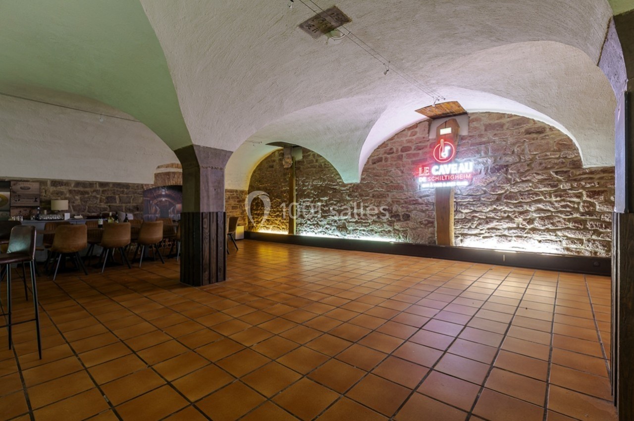 Salle voûtée avec murs en pierre, sol en carrelage orange, chaises alignées et enseigne lumineuse au mur.