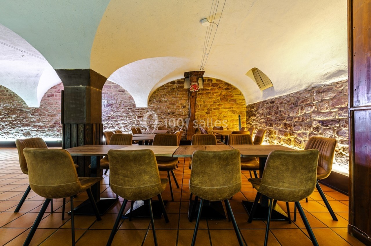 Salle voûtée avec murs en pierre, éclairage chaleureux, tables en bois et chaises rembourrées marron.