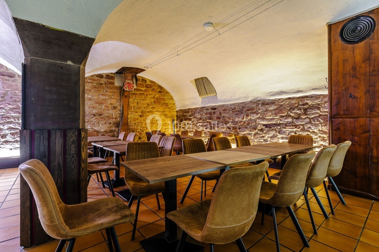 Salle voûtée avec murs en pierre, tables en bois et chaises rembourrées disposées en rangées sur un sol en carreaux.