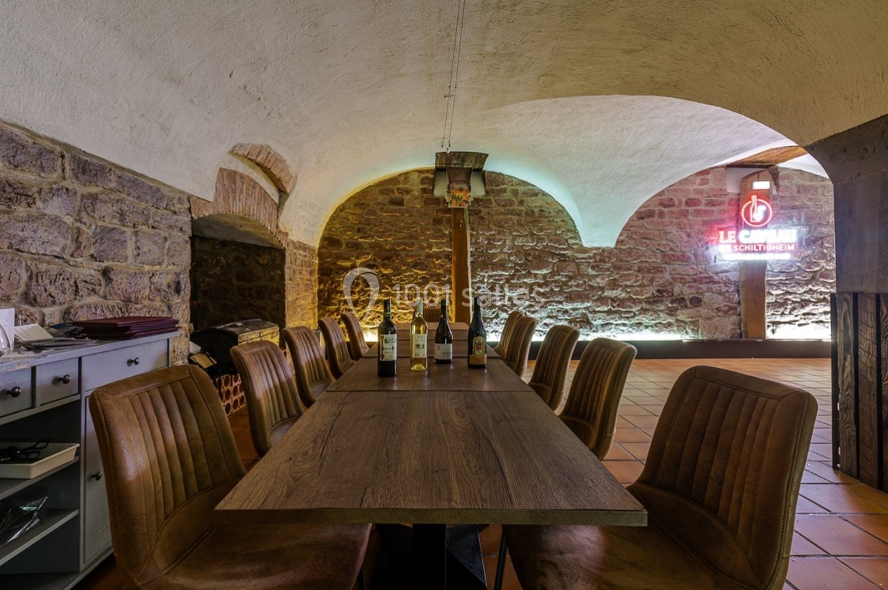 Salle voûtée avec table en bois, chaises rembourrées, bouteilles de vin et mur en pierre éclairé par une enseigne lumineuse.