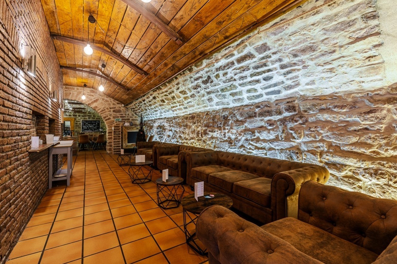 Salle voûtée en pierre avec canapés en cuir, tables basses et éclairage suspendu, sol en carrelage orange.