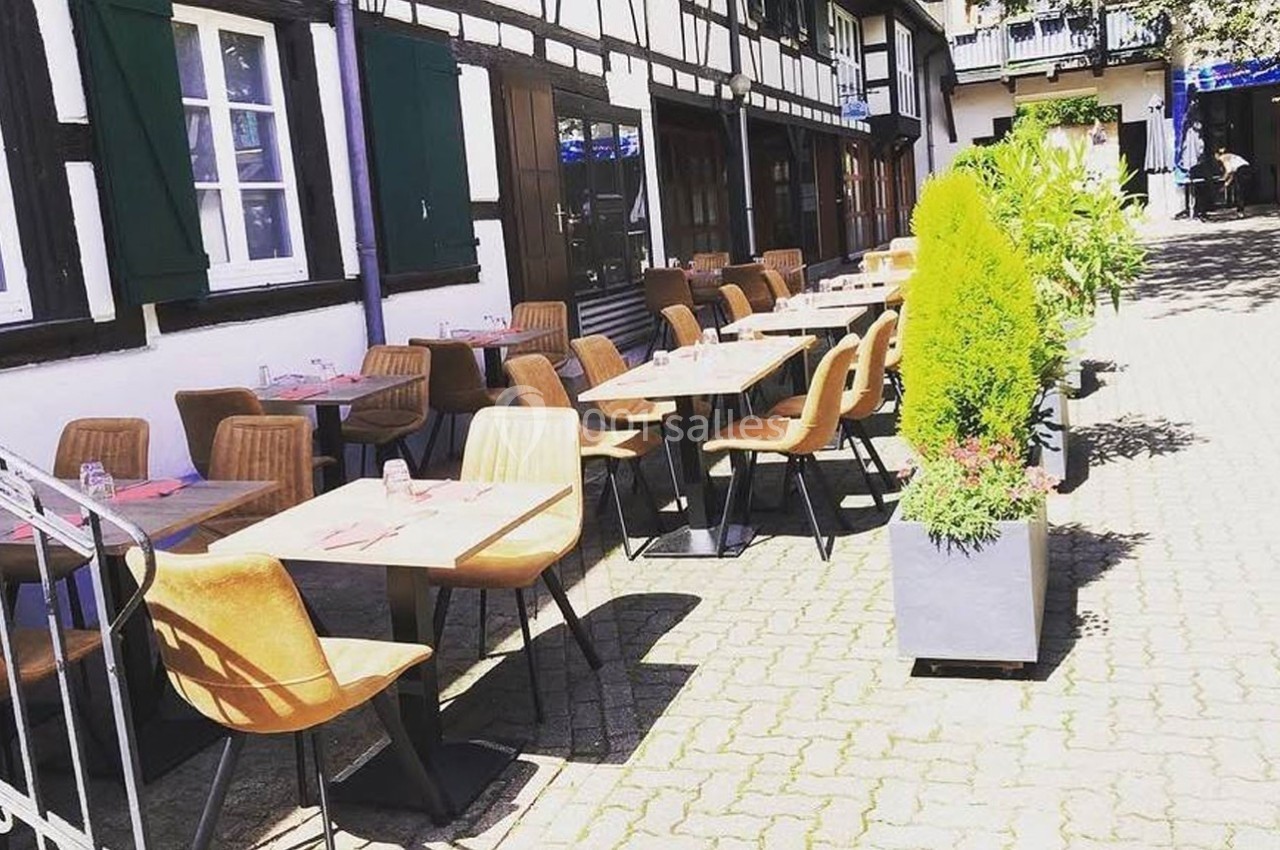 Terrasse extérieure d'un restaurant avec des tables et des chaises alignées, bordée de plantes en pots.
