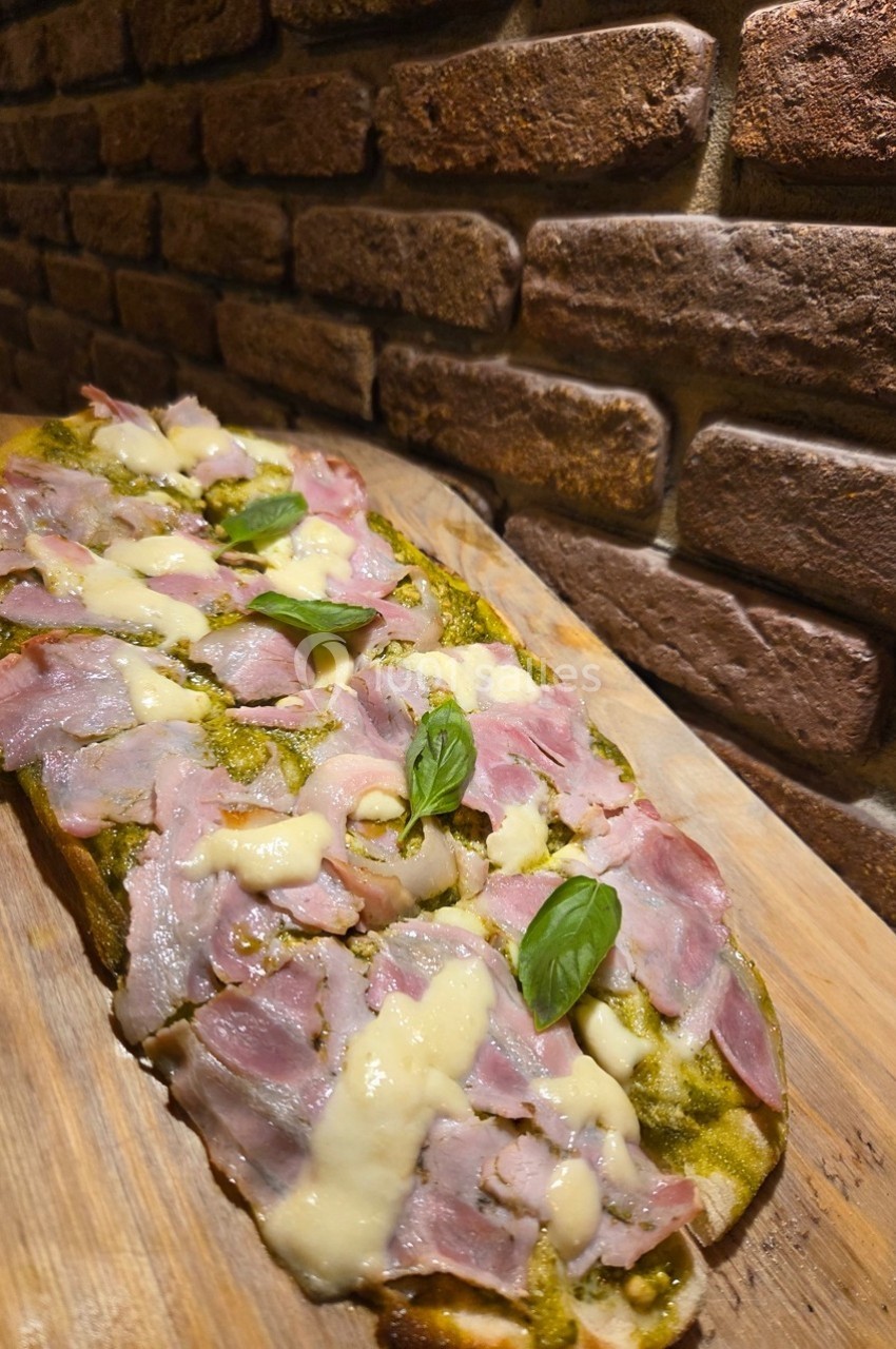 Focaccia garnie de jambon, fromage fondu et pesto, servie sur une planche en bois devant un mur de briques.