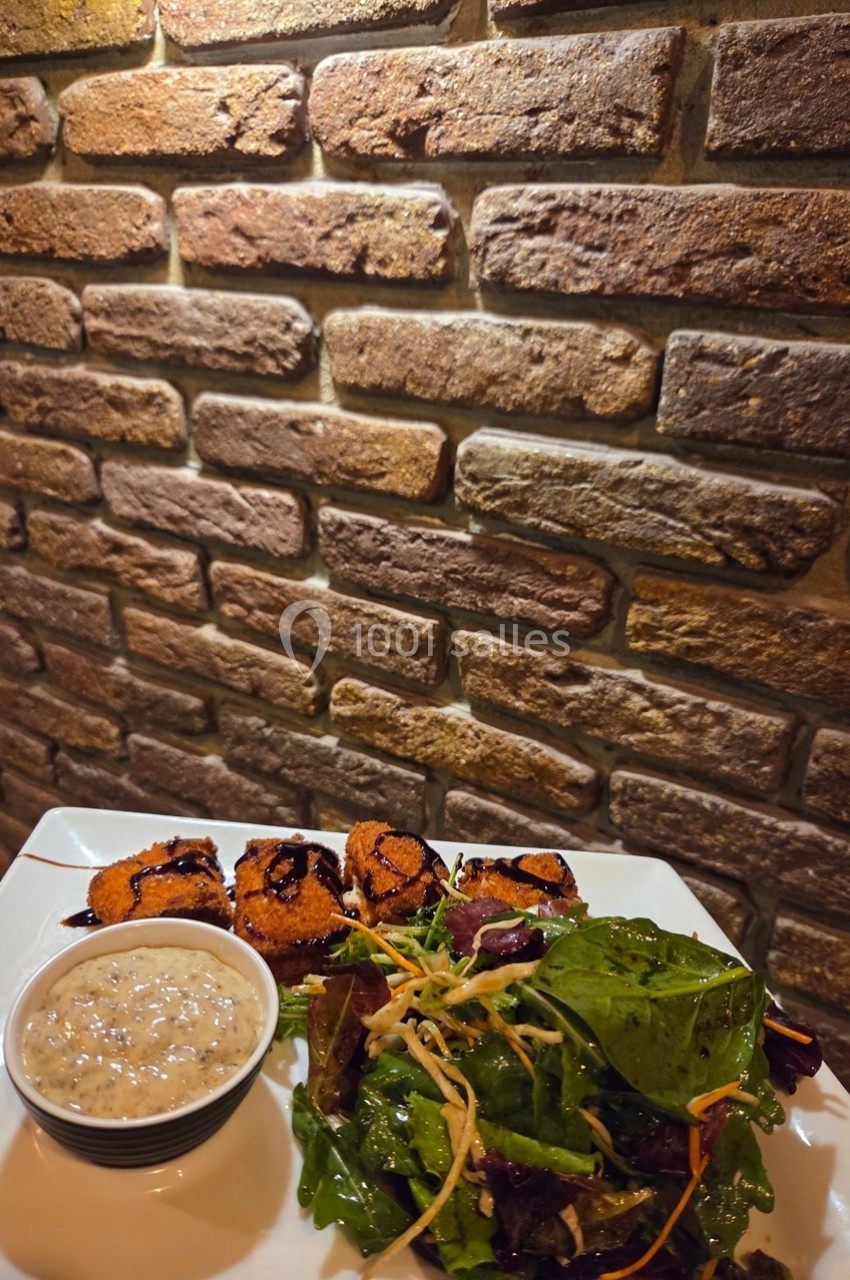 Assiette avec brochettes grillées, salade verte et sauce, posée devant un mur en briques.