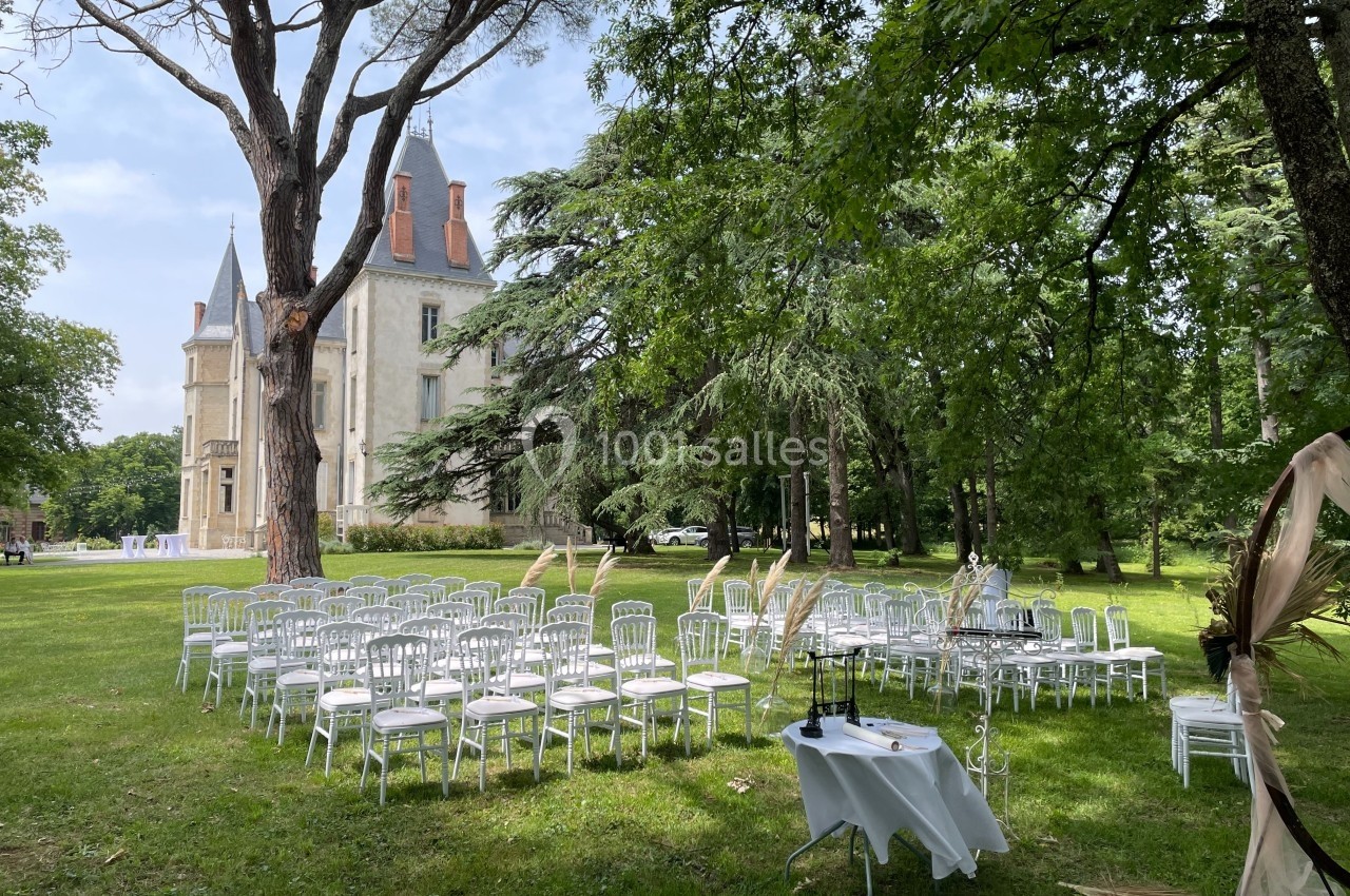 Chaises blanches disposées en extérieur devant un château, entourées d'arbres, pour une cérémonie en plein air.