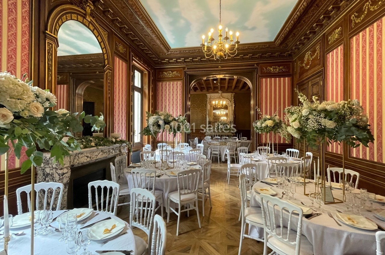 Salle de réception élégante avec tables dressées, chaises blanches, décor floral et boiseries ornées.