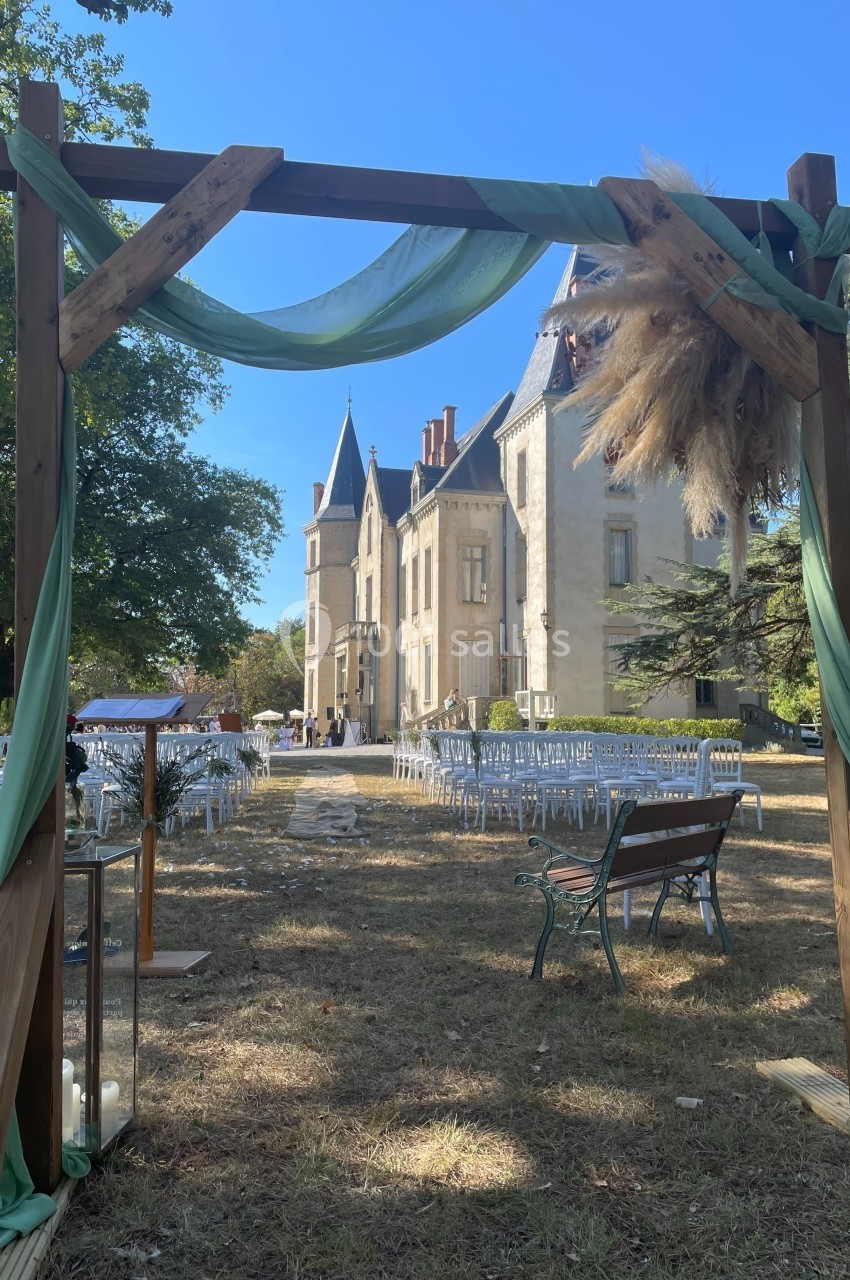 Arche en bois décorée de tissus verts et de fleurs séchées, devant un château avec des chaises disposées pour une cérémonie.
