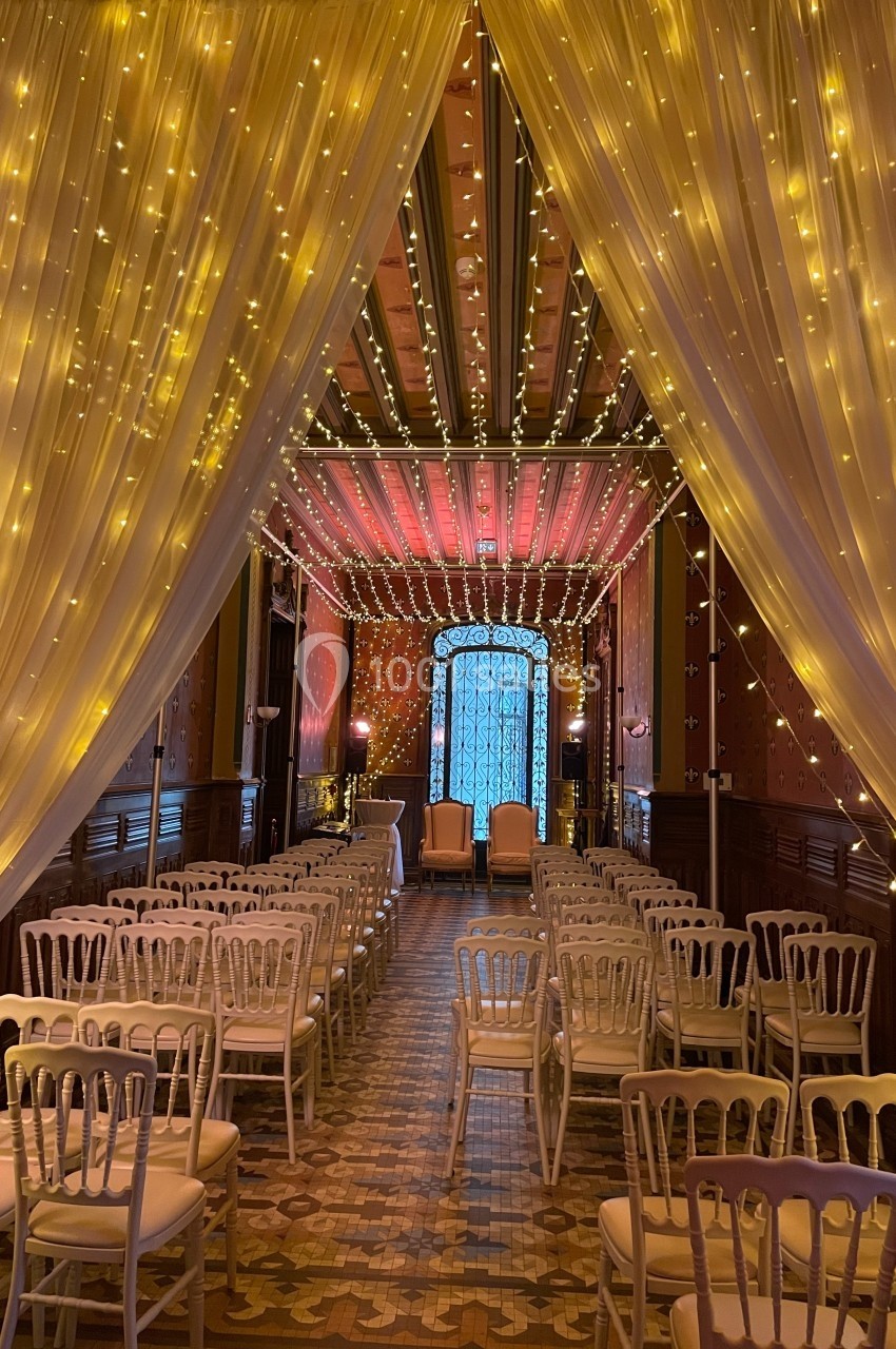 Salle décorée pour un événement avec des chaises blanches alignées, rideaux lumineux et éclairage chaleureux.