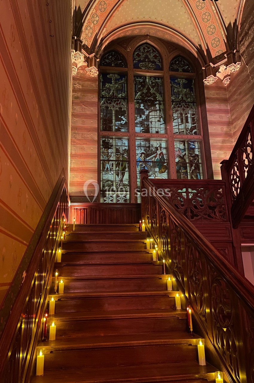 Escalier en bois éclairé par des bougies, menant à un grand vitrail coloré dans un intérieur au style gothique.