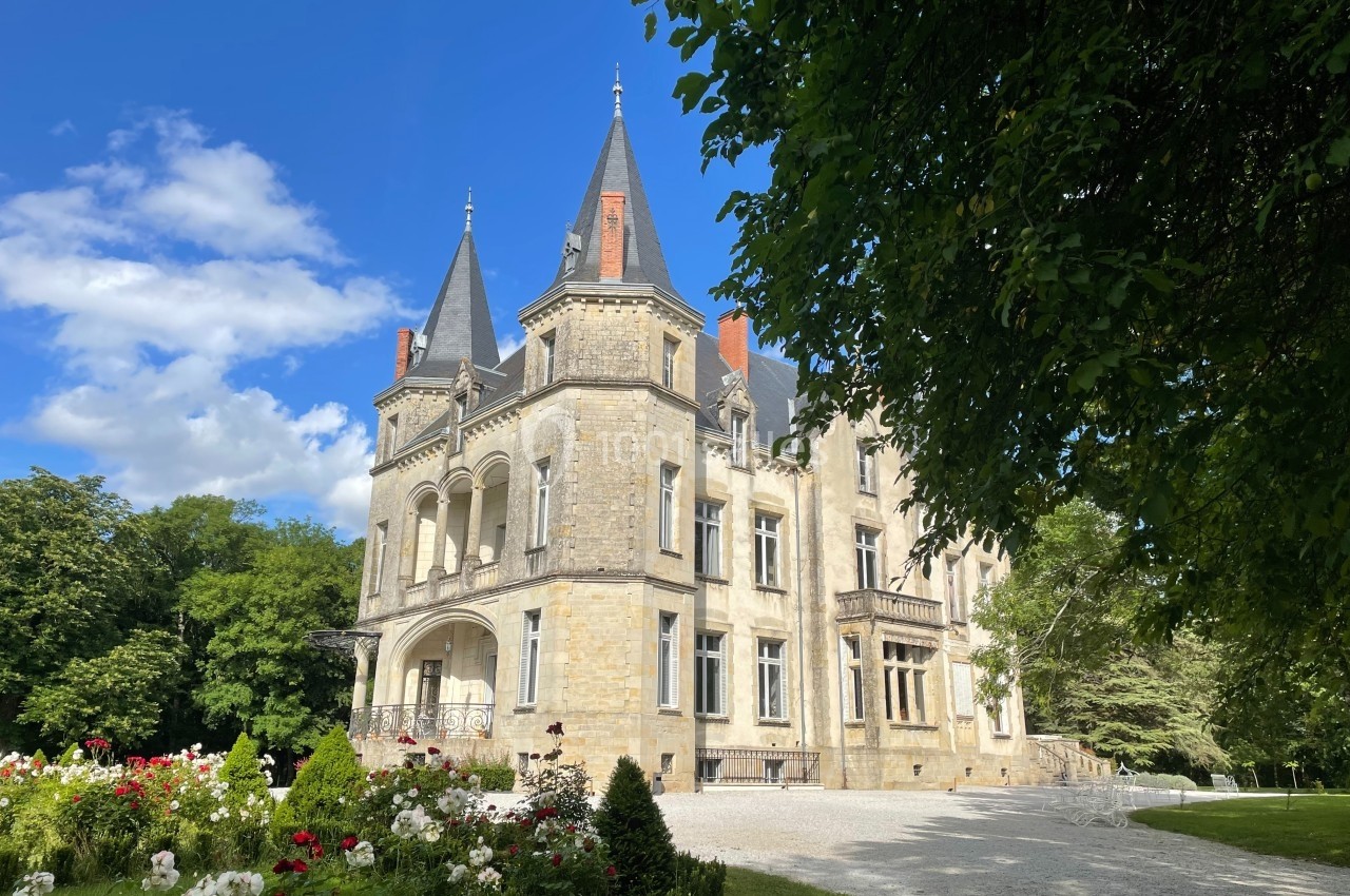 Château en pierre entouré de jardins fleuris, avec un ciel bleu et des arbres en arrière-plan.