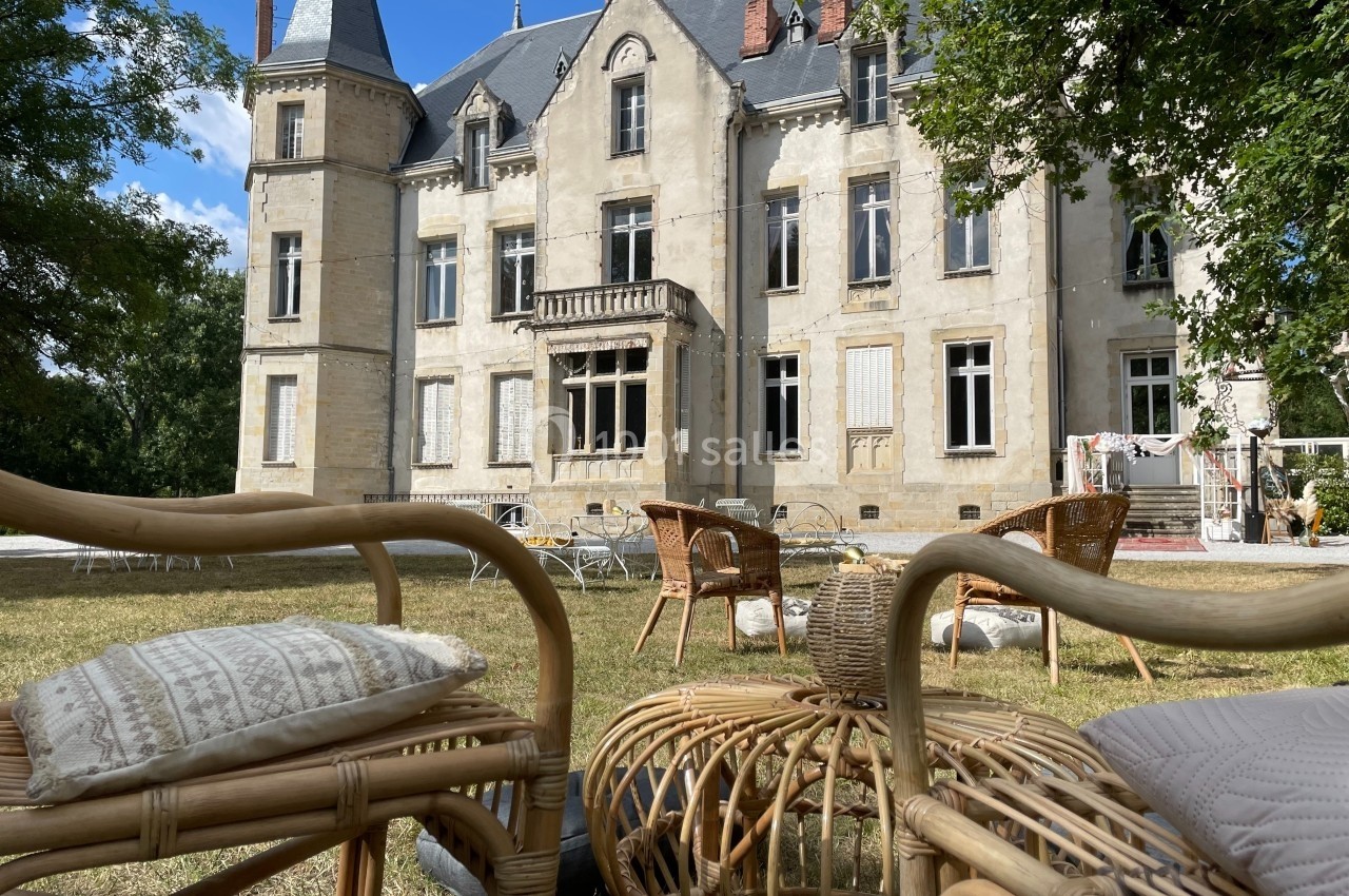 Façade d'un château ancien entouré de verdure, avec des meubles en rotin disposés sur une pelouse au premier plan.