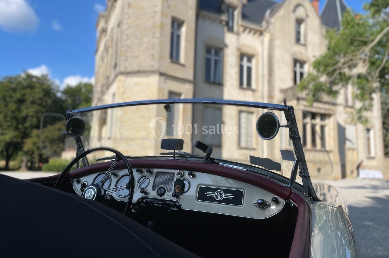 Vue intérieure d'une voiture ancienne décapotable, avec un manoir en pierre en arrière-plan sous un ciel bleu.