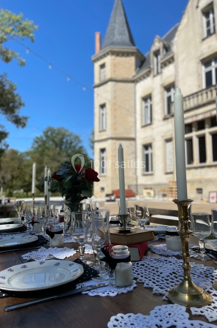 Table dressée avec nappes en dentelle, chandeliers et roses rouges, devant un château sous un ciel bleu.