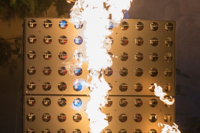 Un homme de dos observe un mur de dispositifs avec des flammes jaillissant à l'extérieur, entouré de végétation.