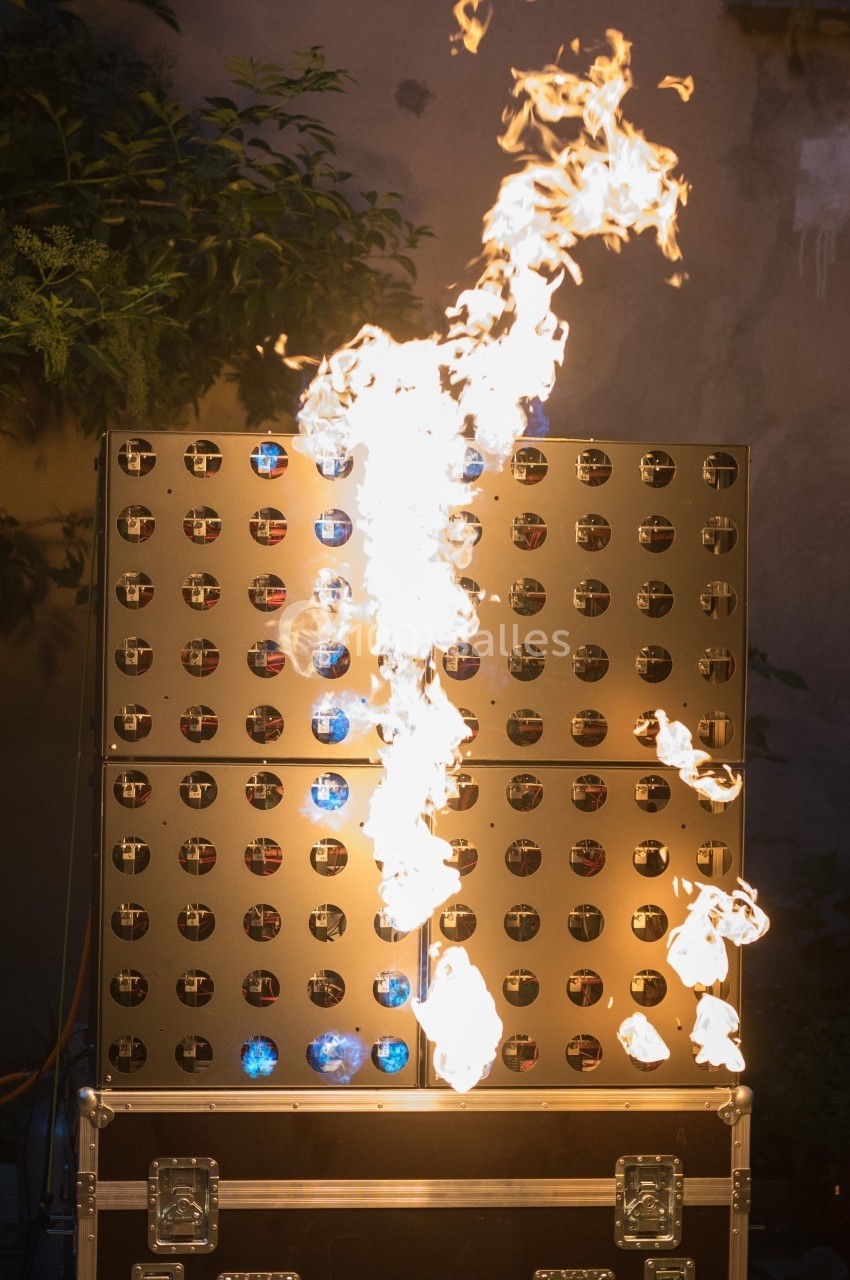 Flammes jaillissant d'un dispositif lumineux en métal perforé, installé en extérieur près de végétation.