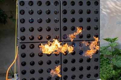 Un homme de dos observe un mur de dispositifs avec des flammes jaillissant à l'extérieur, entouré de végétation.