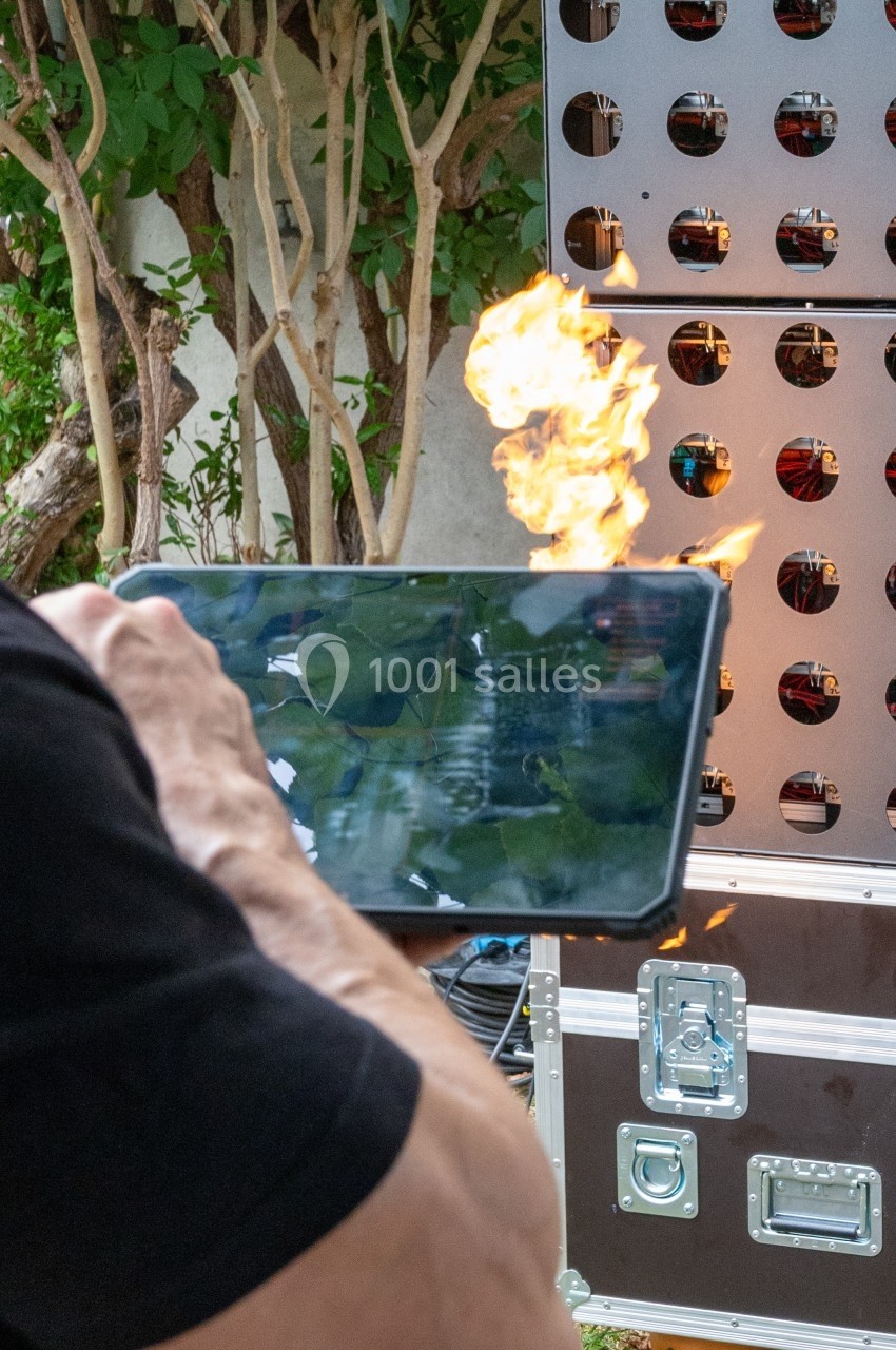 Une tablette tenue devant un mur perforé, avec une flamme visible à l'arrière-plan.